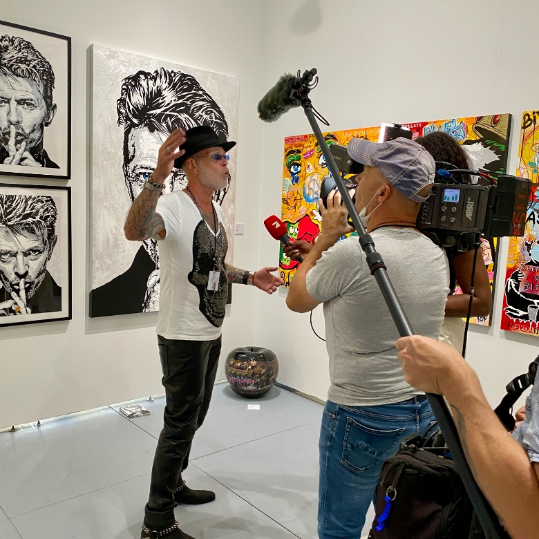 Interview at Scope Art Fair 2021 - Miami Beach Art Basel I wearing a hat and sunglasses gives an interview at Scope Art Fair 2021 during Miami Beach Art Basel, surrounded by pop art and David Bowie-inspired portraits.